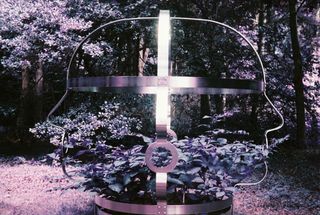 A metal sculpture shaped like the outline of a head with a cross inside it, standing in a purple-tinted forest.