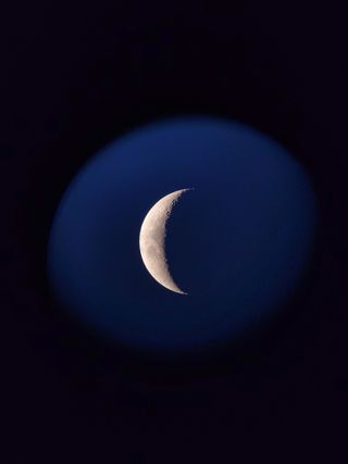 A thin crescent moon inside a round blue glow, against a black sky.