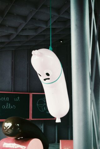 A white inflatable sausage-shaped figure with a sad face hanging from a green cord in a concrete hall, next to a chalkboard.