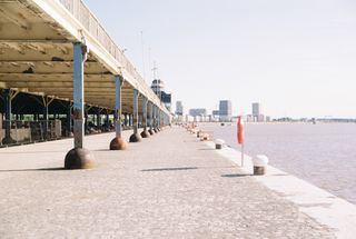 A long covered walkway with iron pillars along a wide river, towers on the horizon.