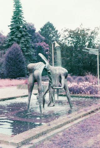 A bronze sculpture of two thin figures in a square fountain, surrounded by pink and purple trees and shrubs.