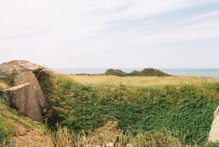 A grassy hollow with chunks of broken concrete, the sea faint on the horizon.