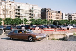 A brown vintage Citroën coupé parked on cobbles in front of a car park and apartment buildings.